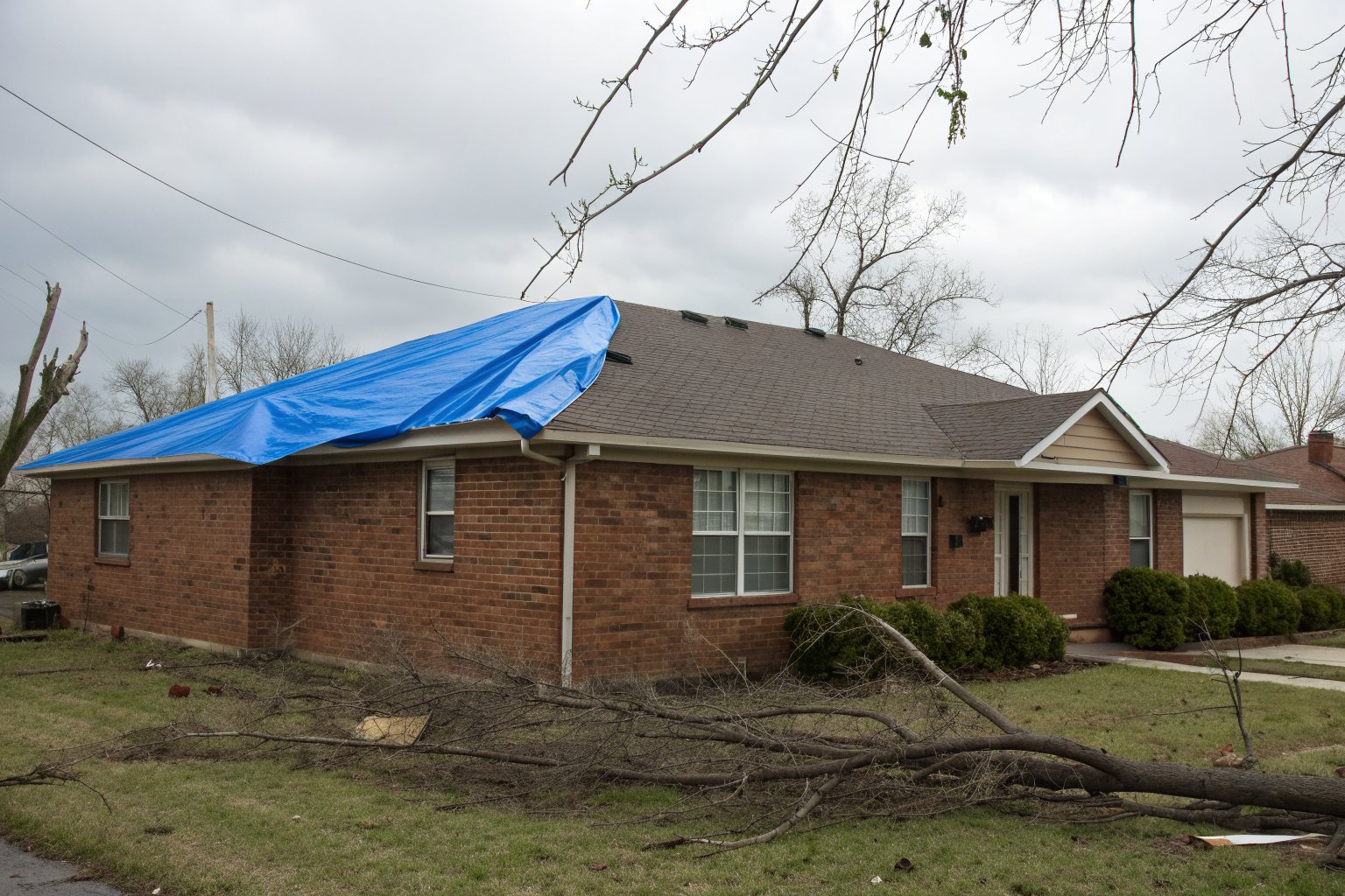 A blue tarp over part of a brick ranch house roof with fallen branches in the yard